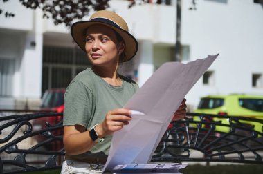 Yeşil gömlek ve hasır şapka giyen bir kadın açık havada bir bankta oturuyor, parlak güneşli bir gökyüzünün altında büyük bir kağıdı inceliyor. Mekan sakin ve çekici bir atmosfer sağlıyor..