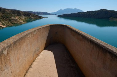 Parlak, berrak bir gökyüzünün altında, arka planda dağlık manzarası olan sakin bir göle bakan beton balkon. Durgun su çevredeki manzarayı yansıtır ve güzel bir manzara yaratır..