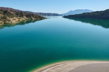 Güzel mavi-yeşil göl, dingin gün ışığında engebeli dağlık kıyı şeritlerini yansıtıyor. Yeşil manzaralar ve sakin sular huzurlu ve resimli bir doğal manzara yaratır..
