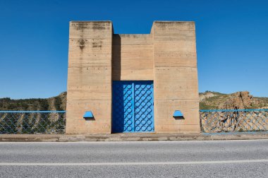 Dairesel geometrik desenlerle bezenmiş parlak mavi bir kapı içeren beton bir yapı, açık mavi gökyüzünün altında eşleşen mavi bir korkulukla tamamlanmış..