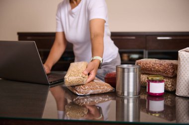 Dizüstü bilgisayar kullanarak, gıda organizasyonu ve dijital planlama kavramlarını tasvir ederken cam bir masada paketlenmiş yiyecek ve konserve ürünlerini sıralayan bir kadın.