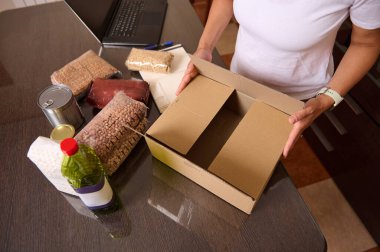 Bir insan, çeşitli temel gıda maddeleriyle çevrili karton bir kutuda erzakları organize eder, bir bakım ve depolama hazırlığı sergiler. Hayırseverlik ya da ev teması için ideal.