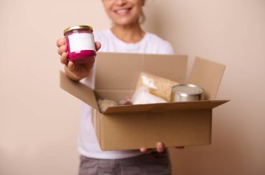 Yemek bankasında elinde karton yiyecek ve içecek kutusuyla gülümseyen gönüllü kadının yakın plan portresi. Kameraya konserve sebzelerle dolu cam bir kutu göstermek. İnsani yardım kavramı