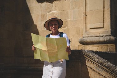 Gülümseyen bir kadın turist tarihi bir şehri ziyaret ederken bir harita üzerinde çalışıyor. Güneş ışığı arka plandaki taş duvarları güçlendirir, sıcak ve davetkar bir atmosfer yaratır..
