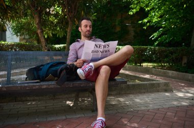 Rahat bir adam rahatça bir bankta uzanır, ağaçların gölgesinde bir gazete okur, huzurlu bir bahçe ortamında, huzur ve rahatlık taşır..