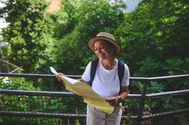 Dışarıda harita okurken gülümseyen bir kadın, güneşin altında yemyeşil bir yer. Keşif, macera, turizm ve doğanın güzelliği için takdiri temsil eder..