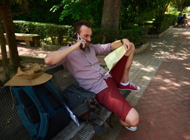 Bir adam parkta bir bankta oturuyor, seyahat planlarıyla meşgul. Etrafı çantalarla çevrili ve günlük kıyafetlerle geziyor. Harita çalışıyor ve telefonuyla konuşuyor. Açık havada güneşli bir günün tadını çıkarıyor..