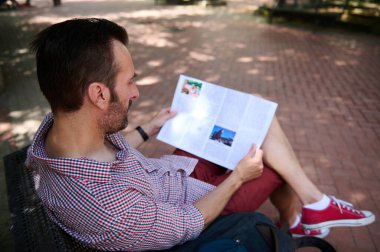 Ekose gömlek ve şort giymiş bir adam bankta oturmuş dergi okuyor ve tuğladan yapılmış zemini ve doğal çevresi olan bir parkta huzurlu bir anın tadını çıkarıyor..