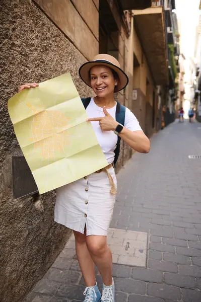 Şapkalı, elinde harita olan ve eski bir şehri keşfeden gülümseyen bir kadın turist. Güneşli bir günde tarihi caddelerde bir seyahat macerasının tadını çıkarıyor..