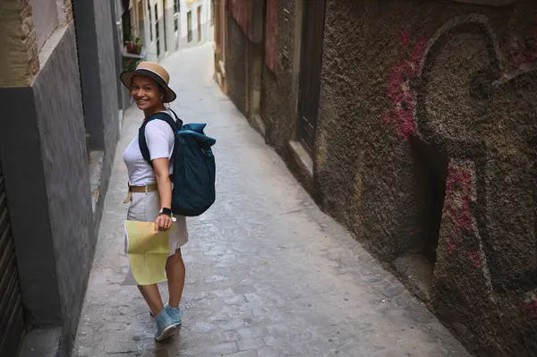 Neşeli bir kadın turist, tarihi bir Avrupa şehrinde, graffiti işaretli dar bir sokakta elinde bir harita ve sırtında bir sırt çantası ile geziyor. Rahat görünüşü seyahatleri ve kültürel keşifleri temsil ediyor..