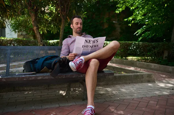 Rahat bir adam rahatça bir bankta uzanır, ağaçların gölgesinde bir gazete okur, huzurlu bir bahçe ortamında, huzur ve rahatlık taşır..