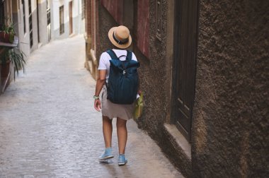 Sırt çantası ve şapkası olan bir kadın eski bir kasabada tuhaf, dar bir sokakta geziniyor. Macera, kültür keşfi ve huzurlu seyahat maceralarını çağrıştırıyor..
