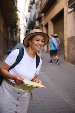 Yaz kıyafetleri ve hasır şapkalı bir kadın eski şehir sokaklarını keşfediyor, elinde bir harita tutuyor, güneşli bir günde yerel atmosferin ve mimarinin tadını çıkarıyor..