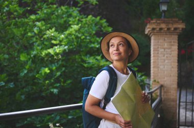 Şapka takan ve sırt çantası taşıyan bir kadın haritayla tarihi bir yeri geziyor. Yeşillik ve mimari unsurlarla çevrelenmiş, merak ve keşif sergiliyor..