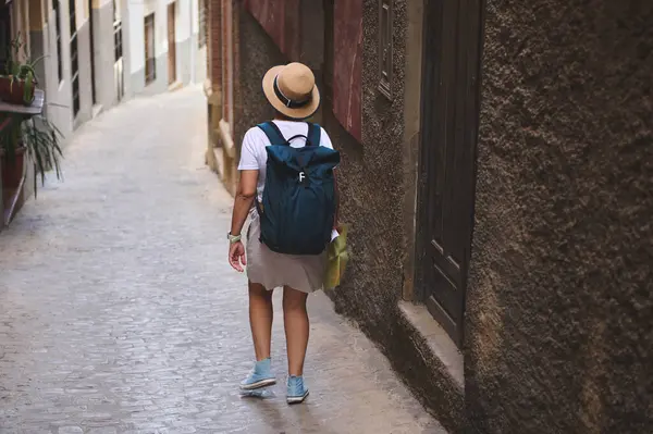 Sırt çantası ve şapkası olan bir kadın eski bir kasabada tuhaf, dar bir sokakta geziniyor. Macera, kültür keşfi ve huzurlu seyahat maceralarını çağrıştırıyor..