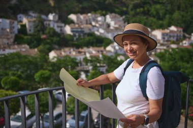 Mutlu bir gezgin elinde bir harita tutarak tarihi bir şehir ortamında, günlük giysiler ve hasır bir şapka giyerek ayakta duruyor..
