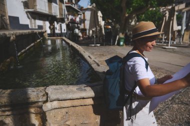 Güneş şapkası ve sırt çantası takan bir kadın harita kullanarak eski ve güzel bir kasabayı keşfediyor. Gün ortası güneşinin tadını çıkarıyor. Bu huzurlu ve resimli ortamda bir taş çeşmenin yanında..