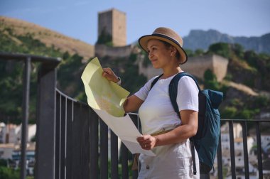 Arka planda etkileyici bir ortaçağ şatosu olan tarihi bir kasabayı gezerken şapka ve sırt çantası takan bir kadın gezgin harita üzerinde çalışıyor. Keşif ve keşiflerin tadını çıkarıyor..