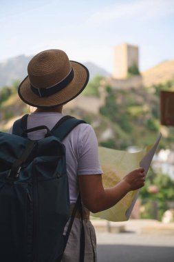 Hasır şapkalı ve sırt çantalı bir kadın, güneşli bir günde, resimli manzaralar ve antik mimarinin ortasında bir haritayla, tarihi bir kasabayı keşfediyor..