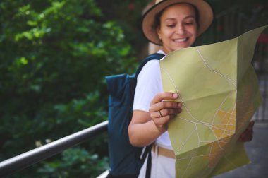 Sırt çantası ve şapkasıyla gülümseyen bir kadın şehir manzaralı bir alanda haritayı inceliyor, keşfetme ve seyahat etme sevincini kucaklıyor..