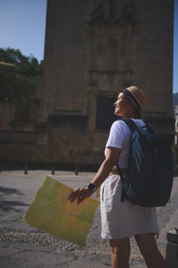Şapka ve sırt çantası takan bir kadın, güneşli gökyüzünün altındaki tarihi bir şehrin mimari detaylarını izlerken bir harita tutuyor..