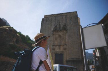Şapka takan ve sırt çantası taşıyan bir turist elinde bir haritayla tarihi bir kasabayı keşfediyor. Çevreleyen mimari ve açık gökyüzü, ortaçağa ait bir yerin cazibesini vurguluyor..