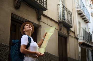 Sırt çantası ve şapkası olan bir bayan gezgin büyüleyici balkonlarla süslenmiş tarihi bir sokakta gezinirken haritayı inceliyor..