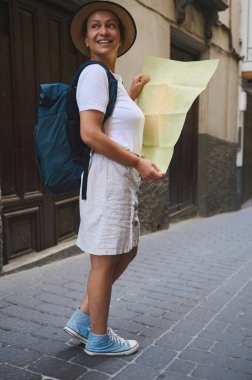 Gündelik yaz kıyafetleri ve sırt çantası giyen bir gezgin, tarihi, eski bir kasabada büyüleyici ve dar bir sokağı keşfederken bir harita tutar, macera ve keşif taşır..