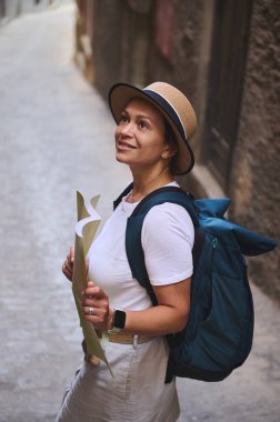 Şapkalı ve sırt çantalı meraklı bir kadın, bir ortaçağ şehrinin dar sokaklarını keşfederken bir harita tutuyor. Resim yolculuk sırasında macera ve keşif taşır..