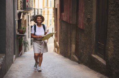 Yaşlı ve güzel bir kasabadan geçerken gülümseyen bir turist kadın. Bir haritayla, güneşli bir günde dar tarihi sokakları keşfediyor, yolculuğun ve keşfin tadını çıkarıyor..