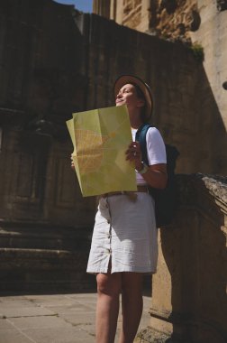 Elinde harita tutan bir kadın, güneşli bir günde tarihi bir şehri keşfetmekten zevk alır. Günlük yaz kıyafetleri ve şapkalar giyer, seyahat ve keşfin ruhunu yansıtır..