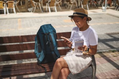 Dişi gezgin, akıllı telefon ve haritaya bakarak, güneşli bir şehir ortamında rahat ve meşgul görünüyor..