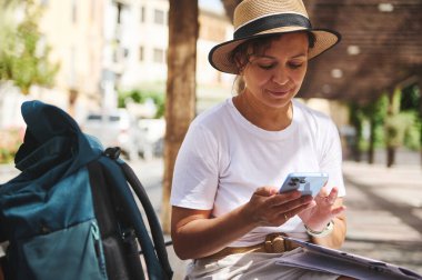 Açık havada oturan, yaz şapkası takan ve günlük kıyafet giyen bir kadın akıllı telefonuyla iletişim kuruyor. Etrafı rahat bir çevreyle çevrili. Bu da seyahat ya da boş zaman aktivitesi olduğunu gösteriyor..