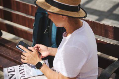 Günlük kıyafetler giyen ve bir şapka takan bayan gezgin akıllı telefonunu kullanarak bankta oturuyor ve yaz günlerinde güneşli havanın tadını çıkarıyor..