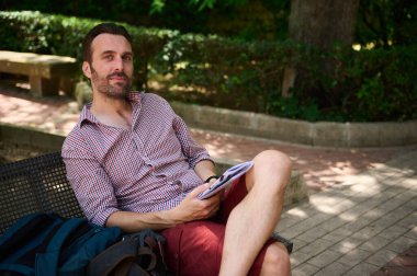 Parktaki bir bankta oturmuş, rahat bir tavırla kitap okuyor, çevresi yeşillik ve barışçıl atmosferle çevrili, gün ışığında dışarıda..