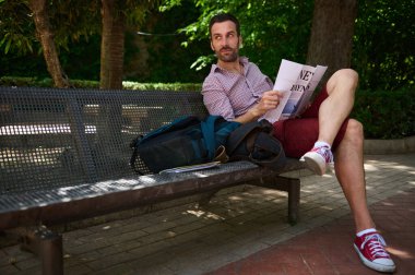 Parktaki bankta oturan bir adam doğal ışık altında gazete okuyor. Yeşilliklerle çevrili, rahat giyimi ve duruşu sakin ve yansıtıcı bir ortam oluşturuyor..