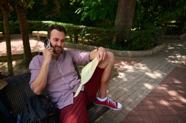 Günlük kıyafetli bir adam, bir parkta bankta oturuyor. Elinde bir harita, telefonda konuşurken yol bulma ve seyahat planlarını aktarıyor..