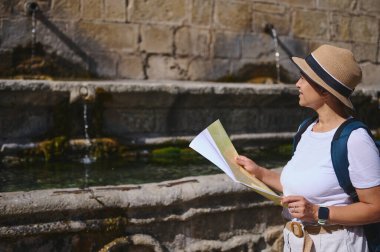 Tarihi bir ortaçağ şehrini keşfeden bir kadın, antik bir fıskiyeye hayran olduğu için elinde bir harita taşıyor. Bir şapka ve sırt çantası takıyor. Macerayı, kültürü ve geçmişteki yolculukları temsil ediyor..