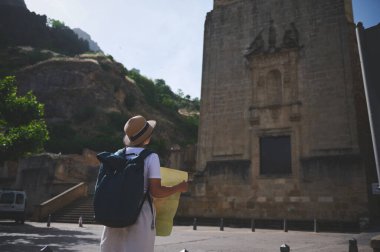 Hasır şapka ve sırt çantası takan gezgin, tarihi bir manzaranın ortasında antik bir taş binanın yakınındaki bir harita üzerinde çalışıyor..