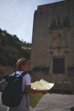 Bir dişi turist, güneşli gökyüzünün altında antik bir kasabanın simgelerini keşfederken haritayı inceliyor, miras ve keşfin cazibesinin tadını çıkarıyor..