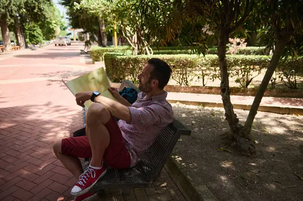 Parktaki bankta oturan bir adam yeşilliklerle çevrili, güneşli bir günde bir harita tutuyor, bir sonraki hedefini planlarken rahatlatıcı ve manzaralı bir deneyimin tadını çıkarıyor..