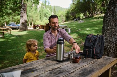 Bir baba, küçük çocuğu açık hava piknik bankında etrafı yemyeşil bir alanla çevrili bir şekilde izlerken eşyalar hazırlar. Görüntü, doğada huzurlu bir aile anını yakalıyor..