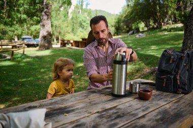 Küçük çocuklu bir adam dışarıda sakin bir anı paylaşıyor. Bereketli yeşilliklerle çevrelenmiş olarak, manzaralı bir günde kaliteli zaman geçirmekten hoşlanırlar..