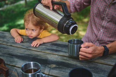 Bir babanın termostan bir bardağa içecek doldurduğu açık hava piknik sahnesi. Genç kızı merakla izliyor, aile bağlarının ve doğa keşiflerinin özünü yakalıyor..