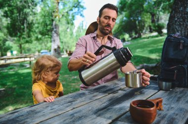 Bir baba çocuğuyla güneşli bir parkta piknik masasında otururken termostan sıcak bir içecek koyar. Sahne, doğada rahat bir aile gezisi olduğunu gösteriyor..