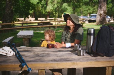 Çocuğuyla birlikte bir piknik masasında oturan bir kadın manzaralı bir orman kamp yerinde, açık havada sükunetin ve aile bağlarının tadını çıkarıyor..
