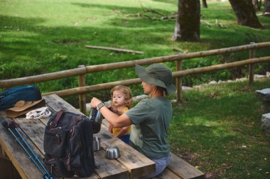 Bir kadın ve çocuk kırsal bir ahşap piknik masasında açık havada aile bağlarını ön plana çıkararak bir anı paylaşıyorlar. Doğa tarafından çevrelenmiş, huzurlu ve ferahlatıcı bir mola veriyorlar..