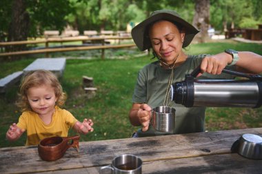 Bir anne termostan sıcak bir içecek dökerken, çocuğu da yeşil bir park ortamında huzurlu bir gezintiye çıkmaya hevesle katılıyor ve açık hava aile anlarının neşesini kucaklıyor..
