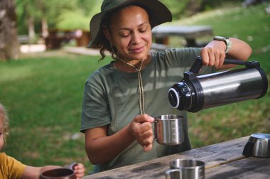 Açık havada huzurlu bir anın tadını çıkaran bir kadın termostan paslanmaz çelik kupaya bir içecek koyar. Çevresi doğanın manzaralı güzelliği ve canlı yeşilliklerle çevrilidir..