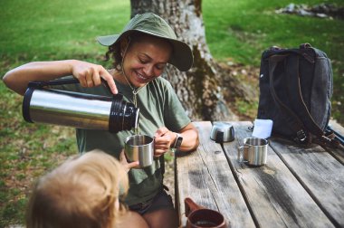 Gülümseyen bir kadın dışarıda oturmuş, rahatlatıcı bir an yaşıyor, yeşilliklerle çevrili ahşap bir piknik masasına çocuğuna içki koyuyor. Doğal ve davetkar bir ortam..